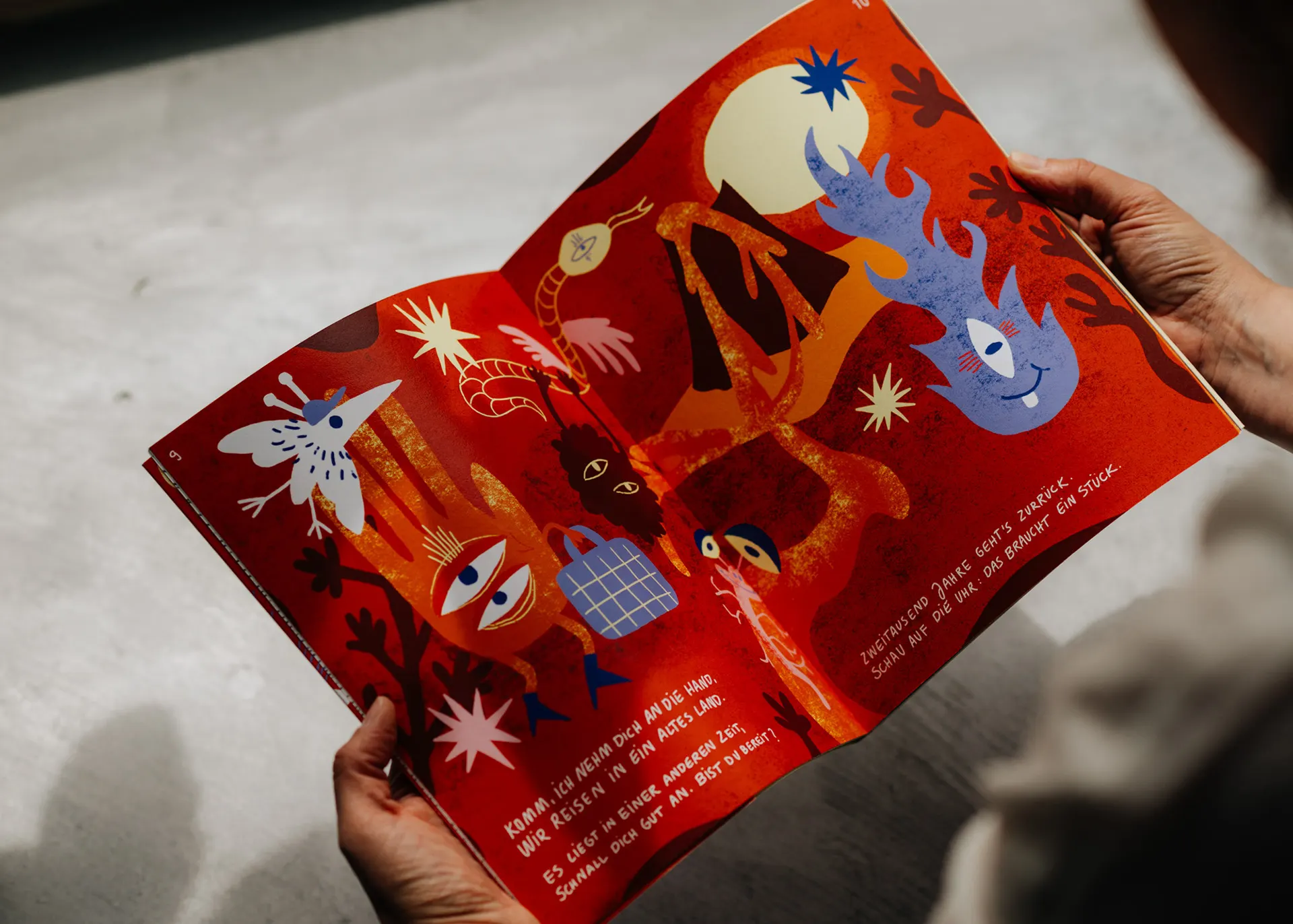 Hands holding the open book, a red spread with figures, flames and stars