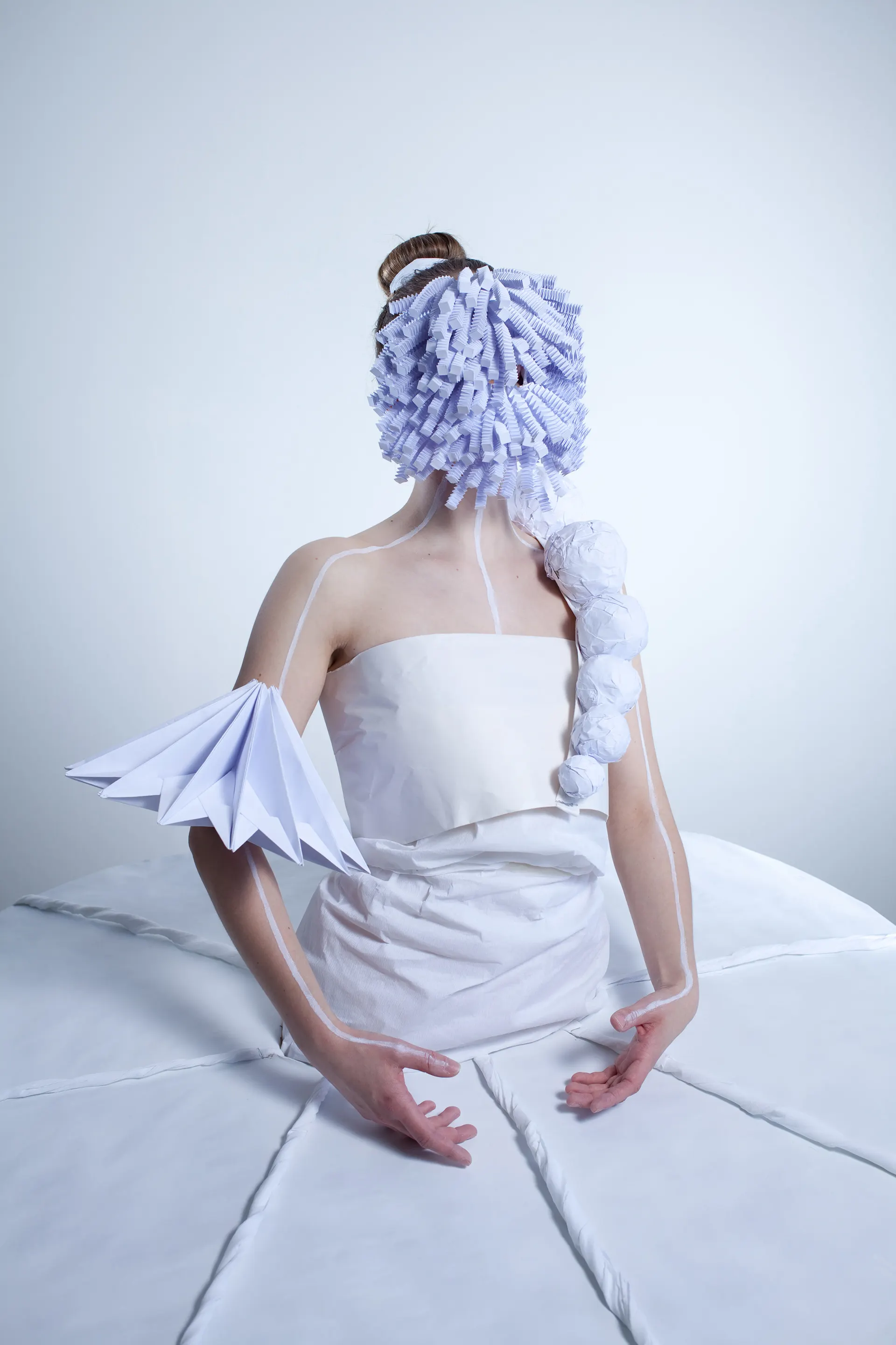 Figure in the paper costume with a mask-like headdress made of paper rolls, against a pale blue background