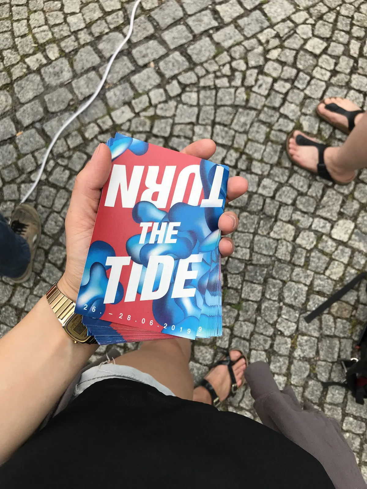 Hand holding a flyer titled “Turn the Tide” above cobblestones