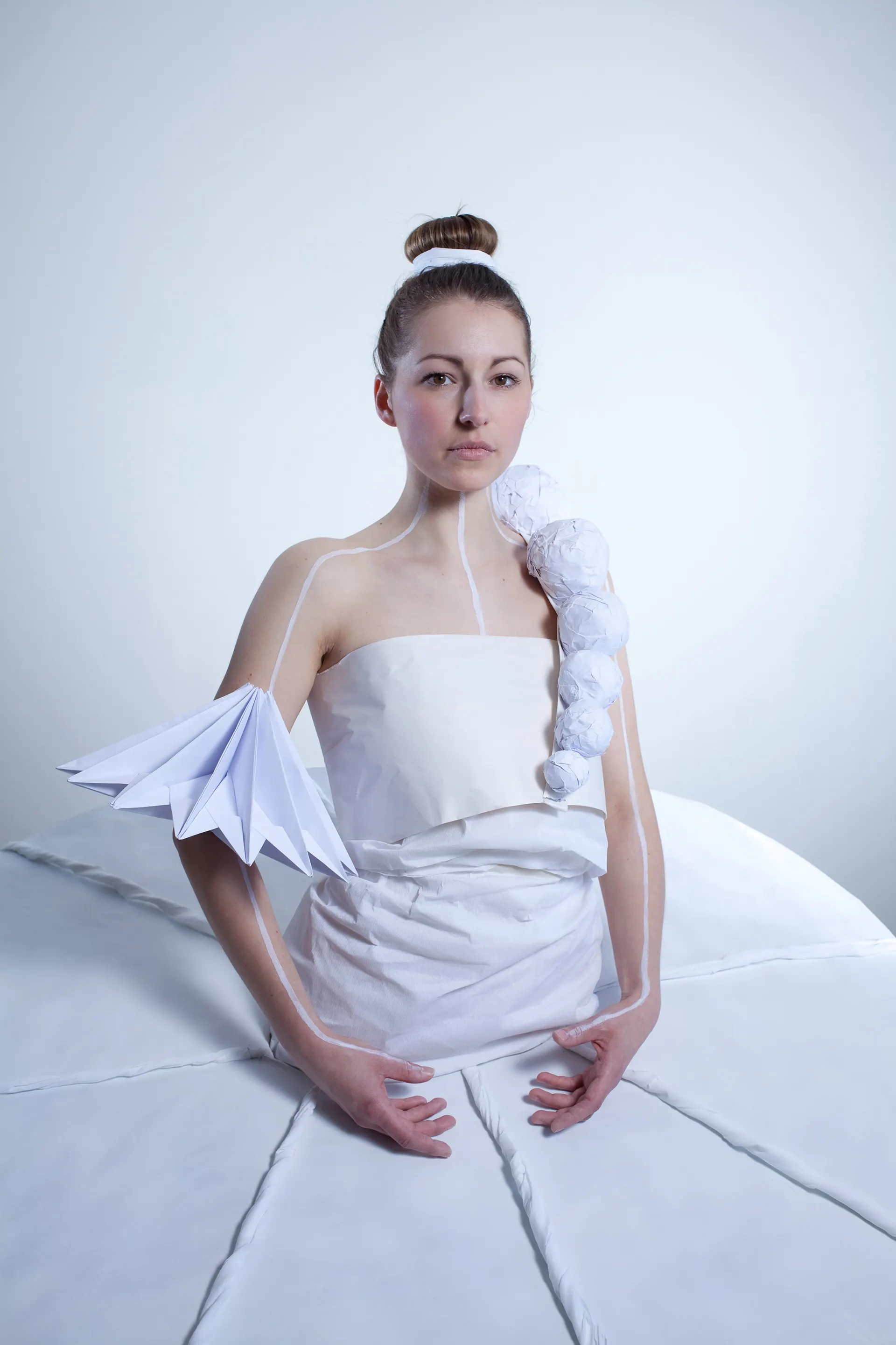 Person in a white paper costume with a folded sleeve and spherical elements on the shoulder, against a pale blue background