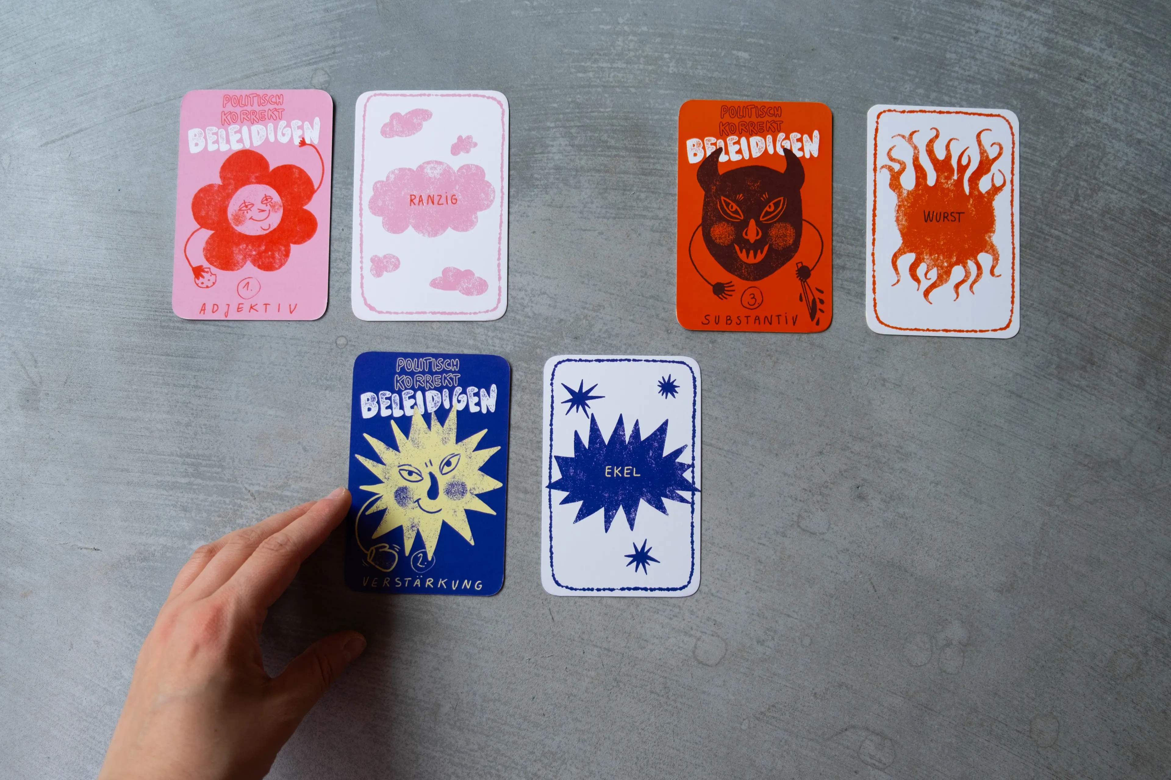 Six cards from the set on a grey surface, a hand placing a card