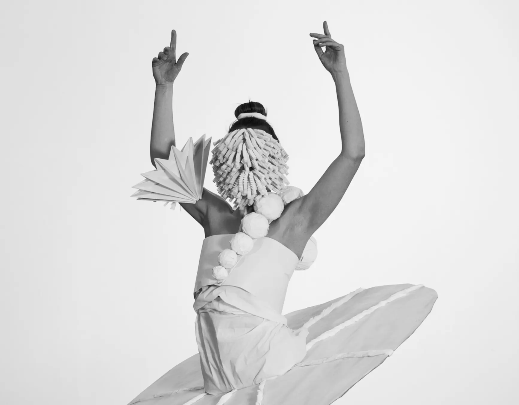 Black-and-white photo of a dancer with raised arms in a paper costume with a fan-shaped sleeve