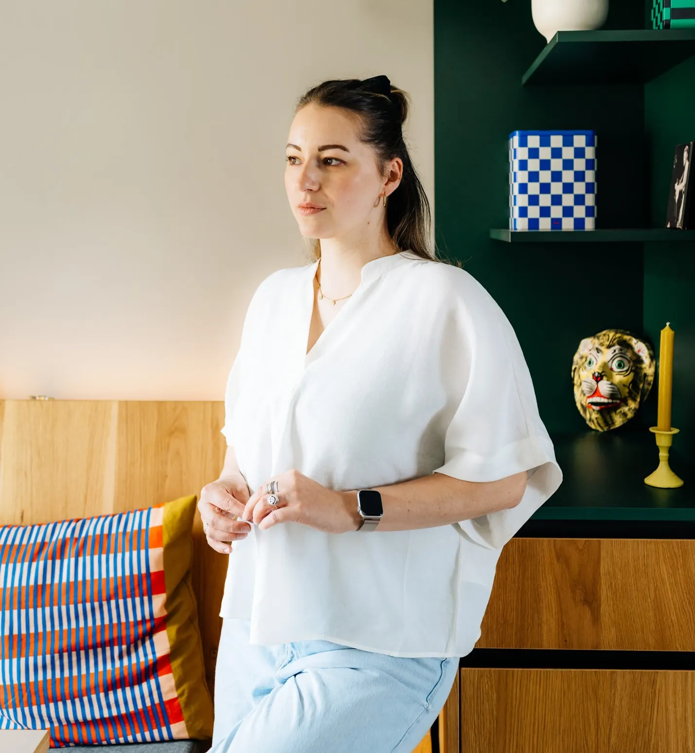 Anne Lutterberg leaning against a wooden cabinet and looking to the side, with a dark green shelf holding a plant and colourful objects behind