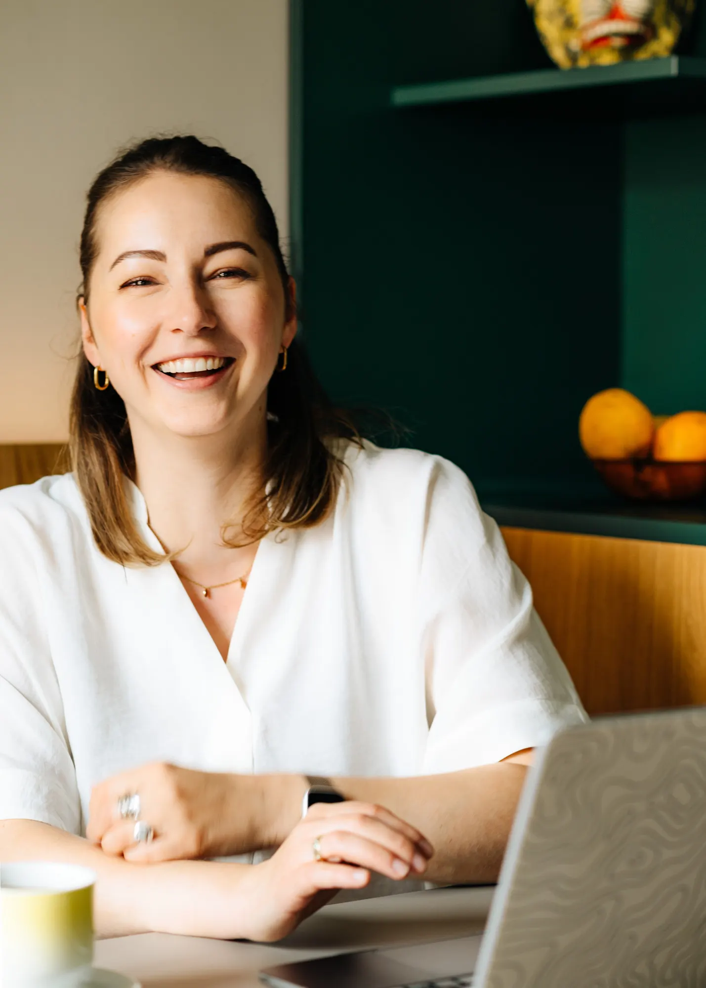 Anne Lutterberg sitting at a table with a laptop and a cup, laughing, with a dark green shelf in the background