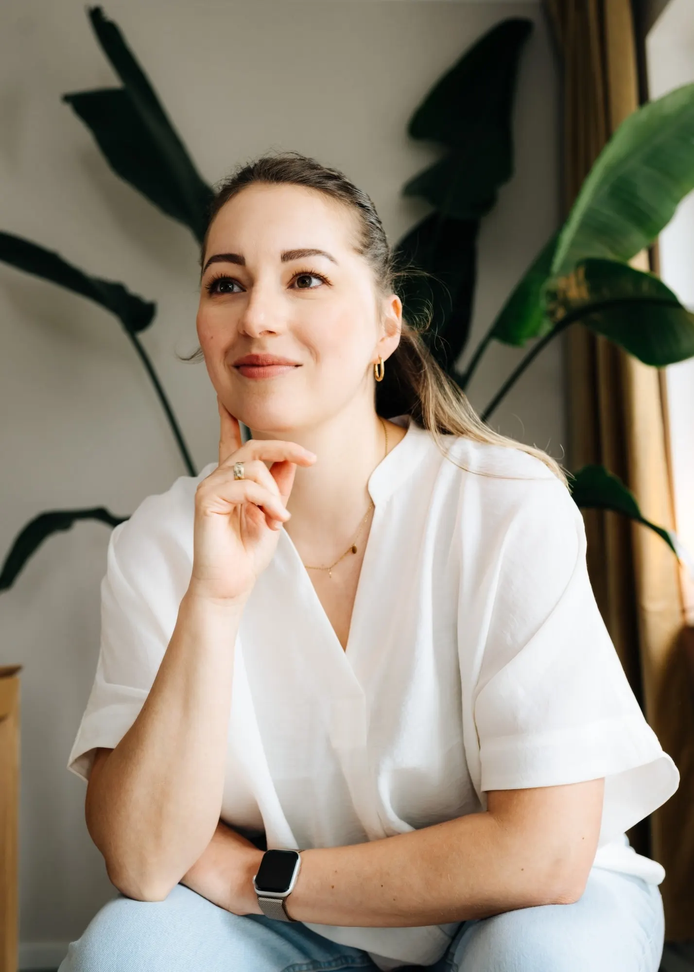 Anne Lutterberg in a white blouse, chin resting on one hand, in front of large plant leaves
